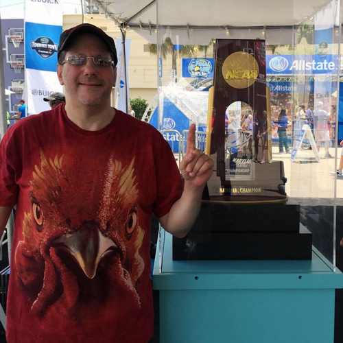 a man holding up his right forefinger stands next to an NCAA national championship trophy 