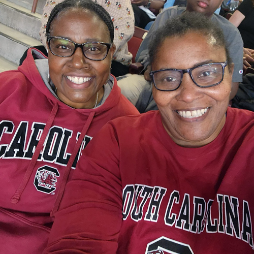 two women wearing garnet south carolina sweatshirts sit in the stands at a ballgame