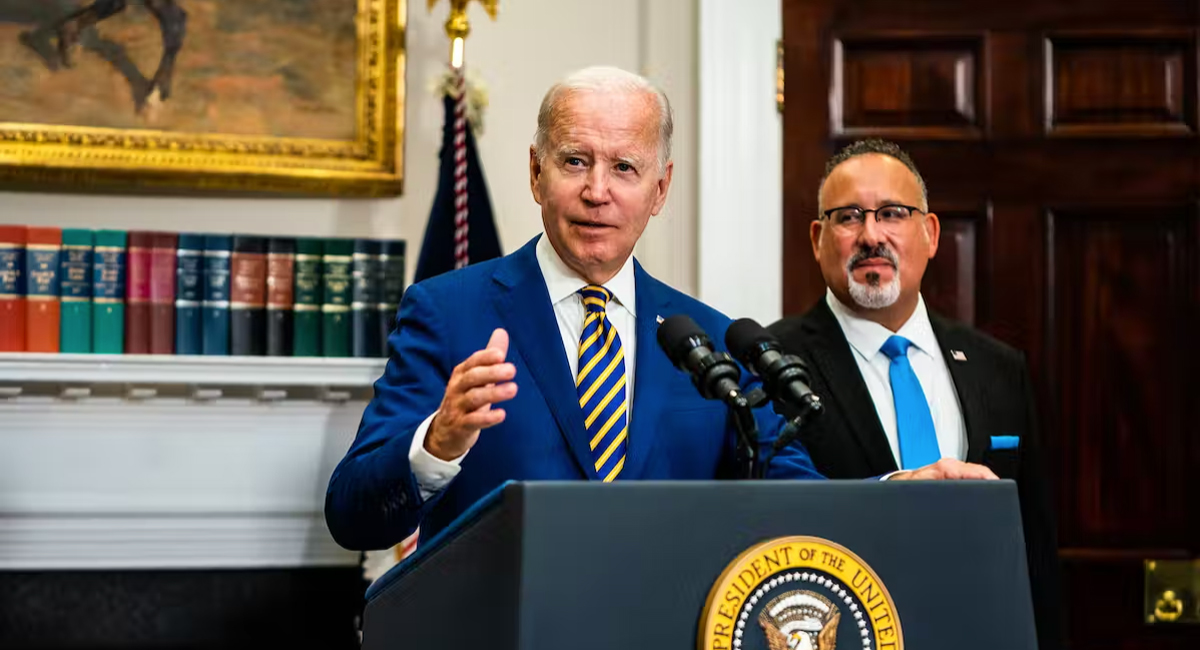 A gray haired man in a royal blue suit jacket with a royal blue and yellow tie, speaks from behind a lectern. A gray bearded, bespectacled man, wearing a black suit and cobalt blue tie, stands behind him and to the left.