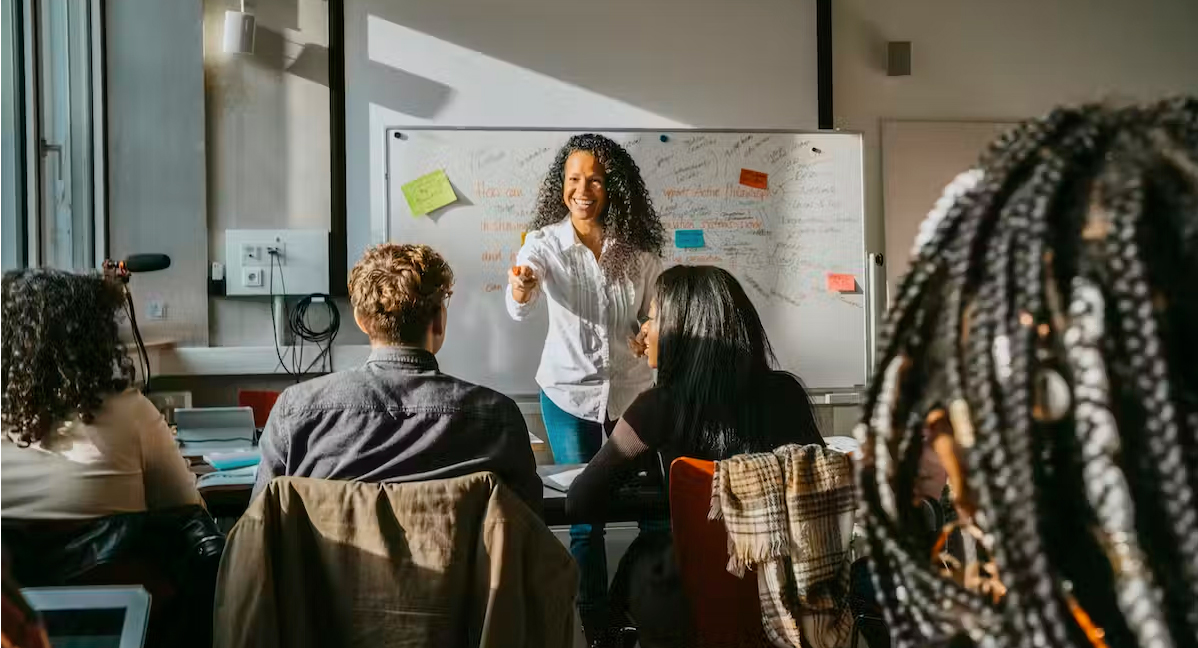 A female teacher stands in front of a whiteboard and faces a group of students