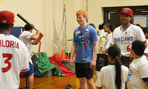 Students of different ages in a gymnasium practicing baseball skills.