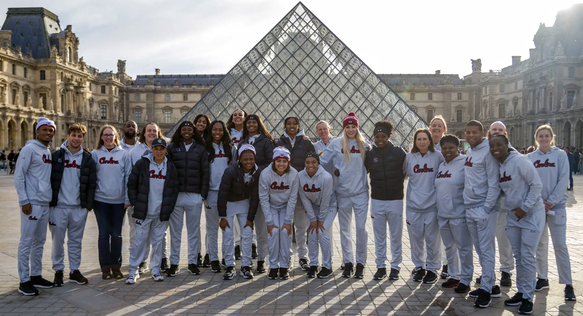 about two dozen people in Carolina sweatshirts pose for a photo outside the Louvre Museum