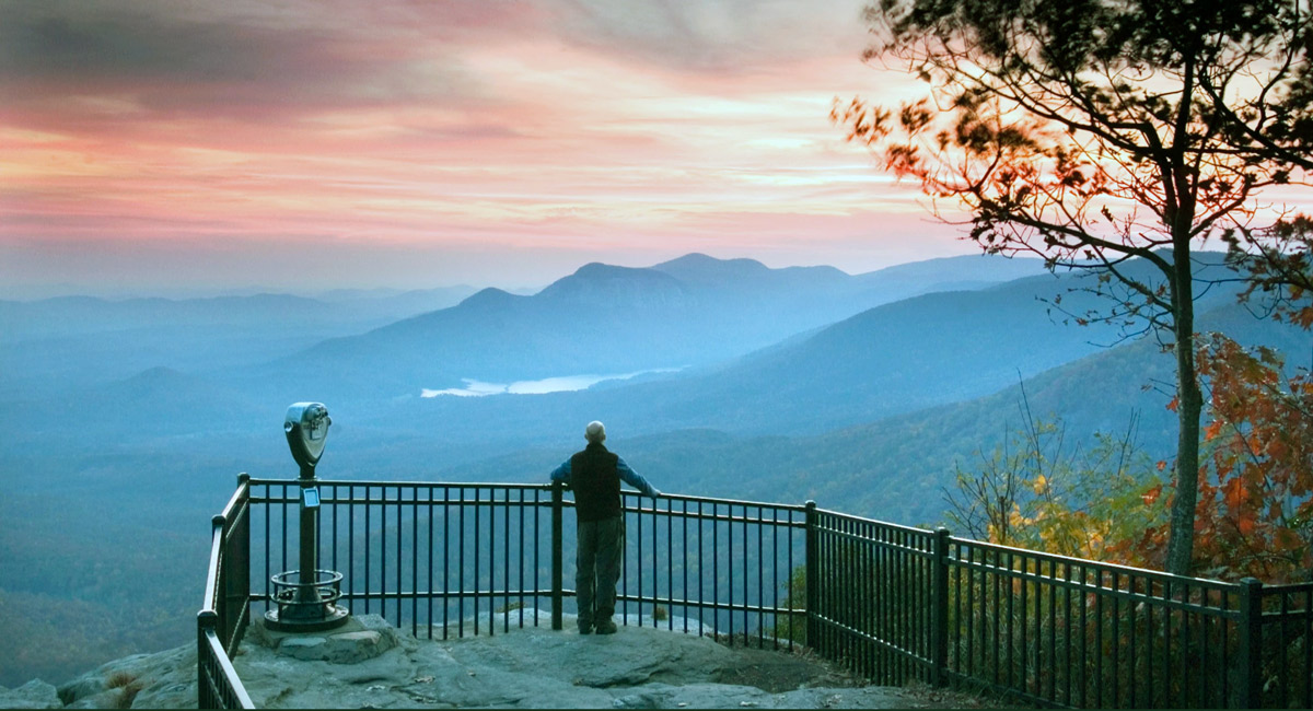 a man stands at a railing looking out on a mountain vista at sunset