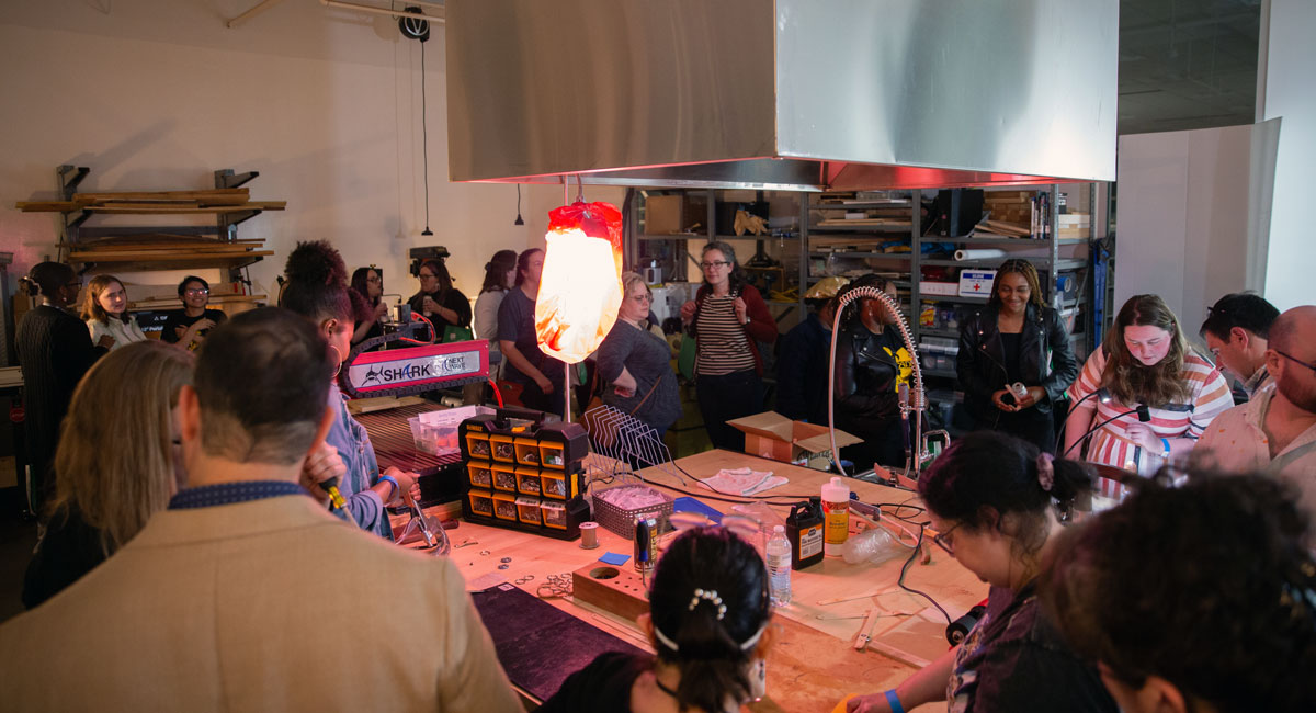 people stand and sit around a table with craft materials and an exhaust fan on the ceiling 