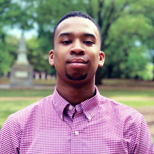 man stands outside with greenery and a monument in the background