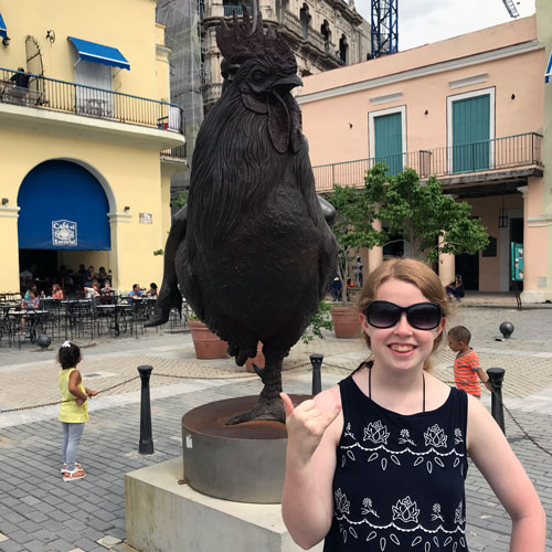 woman stands in a town square with a large rooster sculpture in the background
