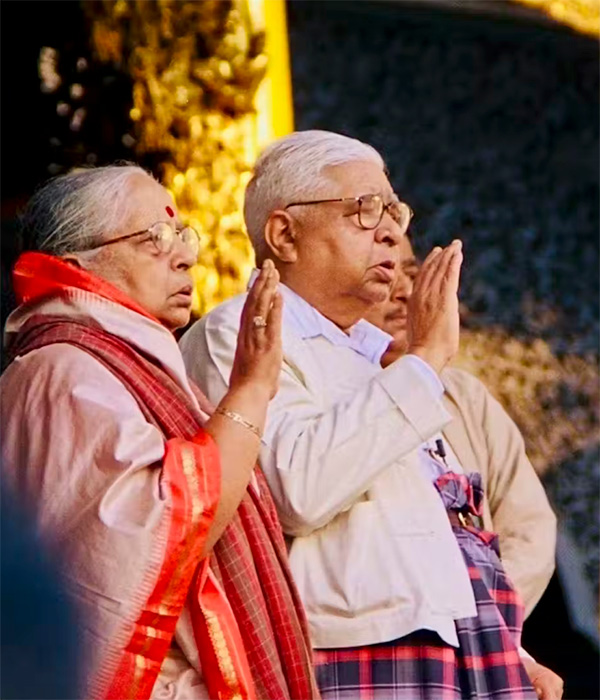 An aging Asian man and woman stand side-by-side with hands held in prayer