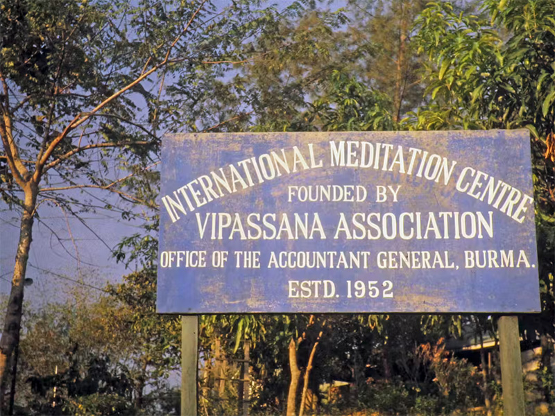 Blue sign with white writing that reads "International Meditation Centre" with wooded area behind