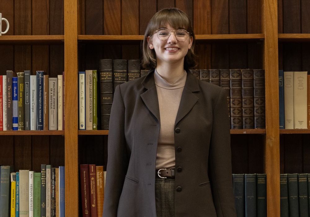 Hannah Keen in brown suit and brown sweater standing in front of bookshelf
