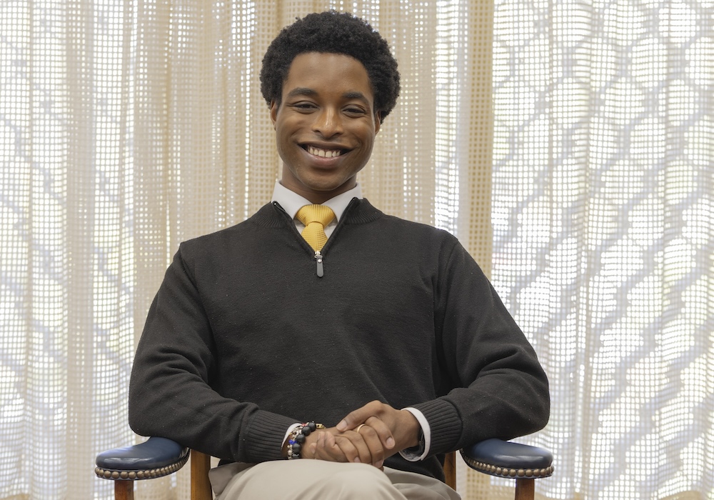 person in a white shirt and yellow tie wearing black pullover sits in a chair in front of a window
