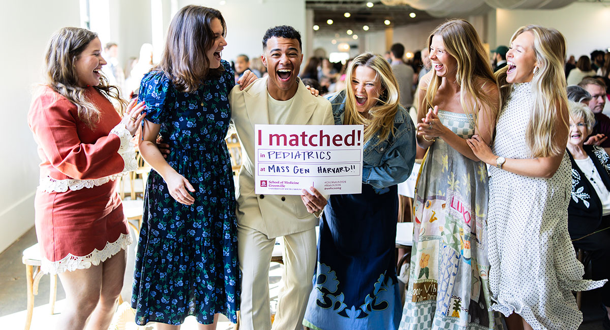 A male School of Medicine Greenville student dressed in a white suite is flanked by five female friends as he holds a sign that indicates he matched for Pediatricss at Mass Gen Harvard.