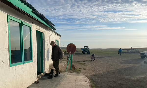 A man silhouetted in the setting sun stands in the doorway of his modest, buff-stuccoed home with green trim in barren landscape.