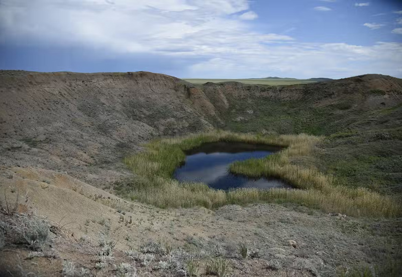 Under blue sky with white clouds, we see some green growth around a crater filled with water, forming a pond, in the valley.