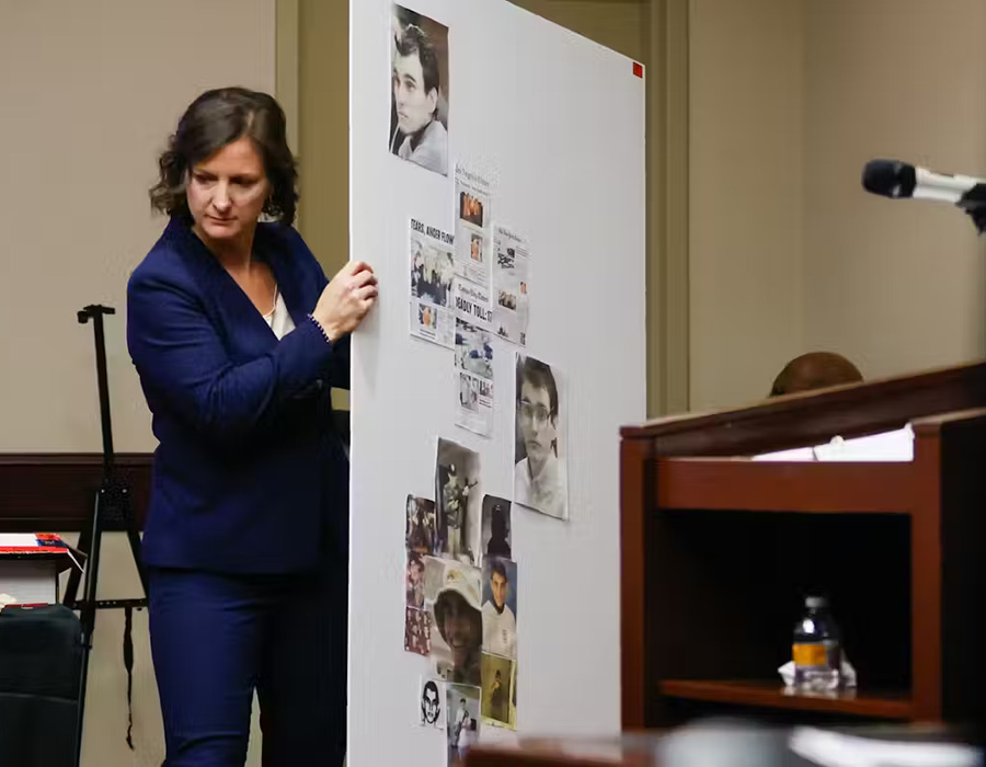 White woman in a dark blue pantsuit holds display board with photo and news clippings to display in courtroom