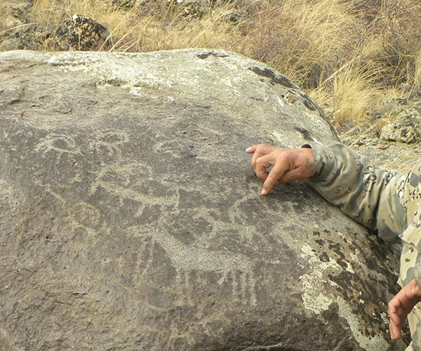 A boulder with researcher's hand pointing to petroglyphs of people and animals.