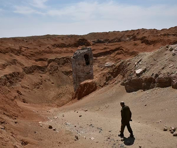 A barren, brown, moonlike landscape with the silhouette of a man in the foreground and excavated silos in the background.