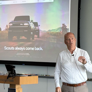 A man teaches in front of a projected slide of a Scout Motors ad.