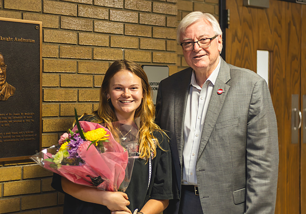 Young woman with shoulder-length brown hair holds flower bouquet standing next to older, suited, gray-haired gentleman