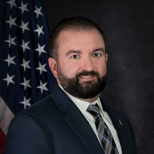 A headshot of Nick Hill in a suit and tie standing in front of an American flag