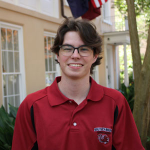 Will Maddux wears a garnet polo and smiles in front of the USC's president's house