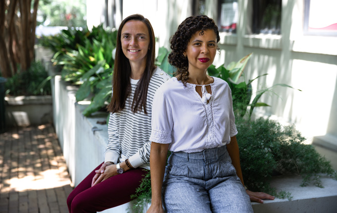 Two College of Education faculty members sit on a concrete ledge in a shaded outdoor corridor surrounded by greenery.
