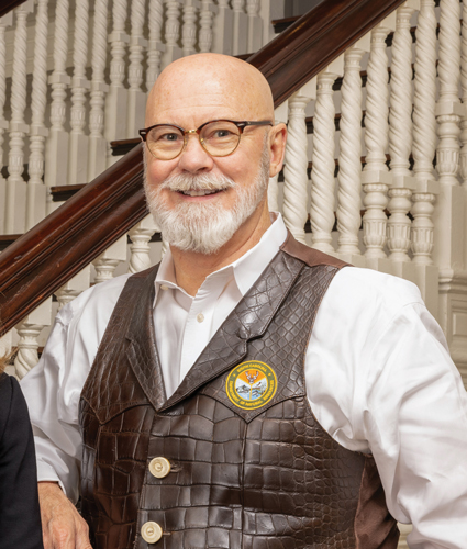 Thomas Mullikins poses in front of a staircase for a portrait wearing glasses and a vest.
