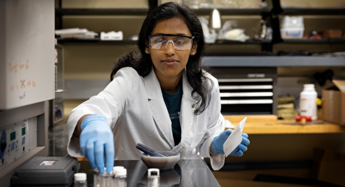 A graduate student wearing protective lab gear prepares materials for an experiment inside a chemistry lab. 