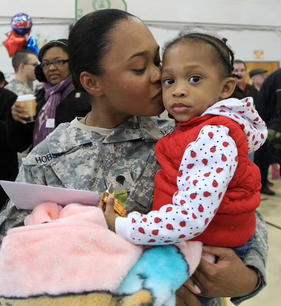 A woman wearing a military uniform holds a small child wearing a red vest and kisses the child's cheek.