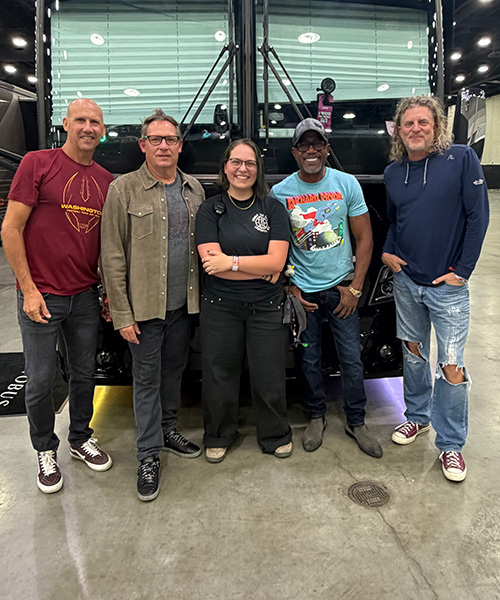 Five informally dressed people pose in front of a band's tour bus