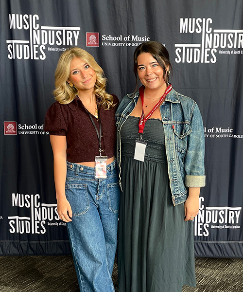 Two women wearing conference badges, blonde in jeans on left, brunette in dress at right, post with industry backdrop