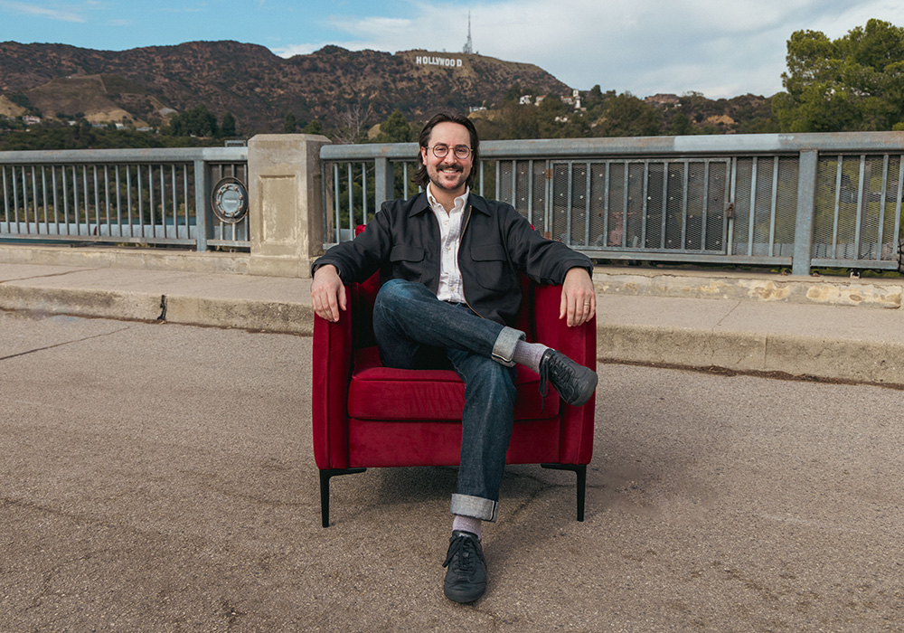 Herman Phillips sits in a red chair outdoors with the iconic Hollywood sign in view behind him.