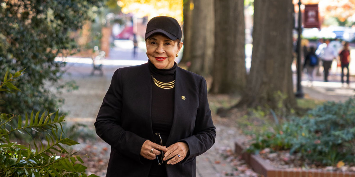 a woman wearing a ball cap stands on a brick walkway with trees in the background